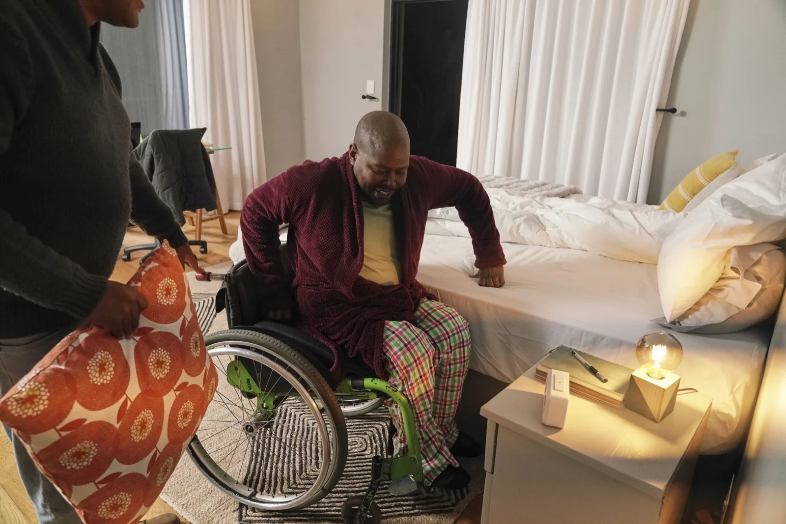 A man moving from his wheelchair onto his bed with support nearby
